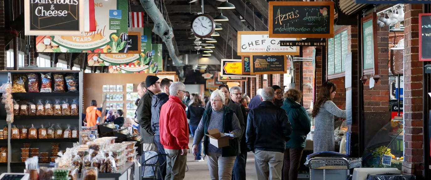 Shoppers walking through Dayton’s 2nd Street Market surrounded by food stalls, artisan vendors, baked goods, and seasonal signage in the historic market hall.