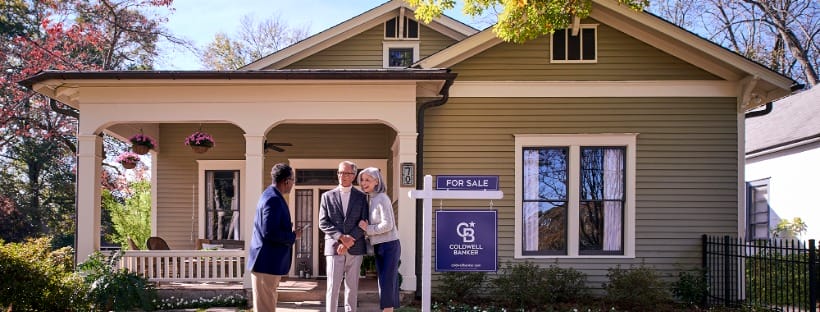 Real estate agent speaking with homeowners on the front porch of a house with a for sale sign.